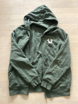 Grön tracksuit True Religion - Säljer hoodien och byxorna tillsammans för 650 men hoodien separat för 400 och byxorna för 250. Storlek L men passar M/L                                         Säljer en grön tracksuit från True Religion med vit logotyp på bröstet och stort tryck på ryggen. Hoodien har dragkedja, ribbade muddar och fickor framtill. Materialet är mjukt och bekvämt, perfekt för chill dagar. Stilen är avslappnad och modern.