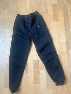 Svarta mjukisbyxor från Polo Ralph Lauren - Säljer ett par svarta mjukisbyxor från Polo Ralph Lauren med klassisk logga broderad på vänster ben. Byxorna har dragsko i midjan och muddar vid bensluten. Perfekta för chill eller träning, tillverkade i mjukt material som känns skönt mot huden.