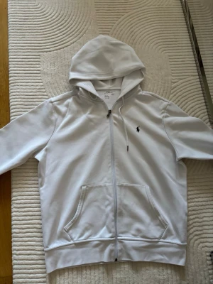Vit hoodie från Polo Ralph Lauren - Snygg vit hoodie med dragkedja från Polo Ralph Lauren. Klassisk design med huva, kängurufickor och den ikoniska lilla loggan broderad på bröstet. Perfekt för en clean och stilren look. Tillverkad i mjuk bomull för extra komfort.