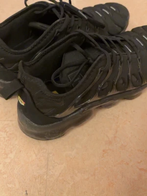 Nike Air Max Plus TN Svarta sneakers - Svarta Nike Air Max Plus TN sneakers med ikonisk vågig design och synliga Air-dämpningar i sulan. Skorna har svarta snören, mesh- och syntetmaterial samt små reflekterande detaljer på ovandelen. Perfekta för dig som gillar sportig och streetinspirerad stil.