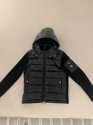 Mörkblå Moncler cardigan - Snygg mörkblå cardigan från Moncler med glansig vadderad front och huva. Ärmarna är i mjukare tyg och har en ficka med Moncler-logga på vänster arm. Jackan har dragkedja framtill och två fickor med tryckknappar. Perfekt inför hösten/våren. Storlek Xl men passar även för dom som har L då Moncler är små i storlekarna . Jackan är äkta och köpt på plick för 5000, kom privat för fler frågor.