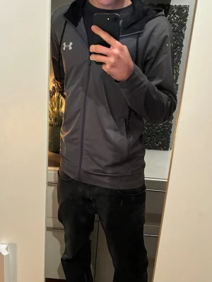Grå hoodie från Under Armour - Snygg grå hoodie från Under Armour med dragkedja och svart huva. Tröjan har två sidofickor med dragkedja och en vit logga på bröstet. Perfekt för dig som gillar sportig och stilren look. I bra skick, inget hål eller någon lös tråd. Inga fläckar. Nypris 399kr 