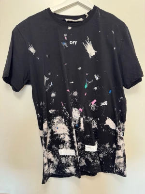  t-shirt med tryck - Cool svart t-shirt fmed vit logga och färgstänk över hela plagget. Stort grafiskt tryck på ryggen och texten 'OFF' framtill. Klassisk passform och rund hals. Perfekt för dig som gillar streetwear och unika detaljer.