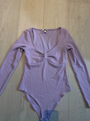 Lila ribbad body med lång ärm - Trendig lila body med lång ärm och djup urringning. Ribbad struktur och twistad detalj framtill vid bysten ger en snygg siluett. Perfekt att styla till jeans eller kjol för en cool look.