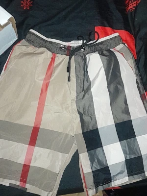 Rutig shorts med snörning - Snygga shorts med klassiskt rutigt mönster i beige, svart, vitt och rött. Shortsen har elastisk midja med snörning för skön passform. Perfekta för sommardagar och avslappnade tillfällen.