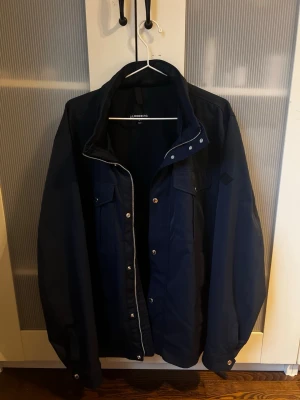 Mörkblå J.Lindeberg jacka  - Snygg mörkblå vindjacka från J.Lindeberg med klassisk krage och knappar framtill. Jackan har flera fickor och en diskret design som passar perfekt till en modern stil. Materialet är troligtvis polyester och jackan har långärmad passform.