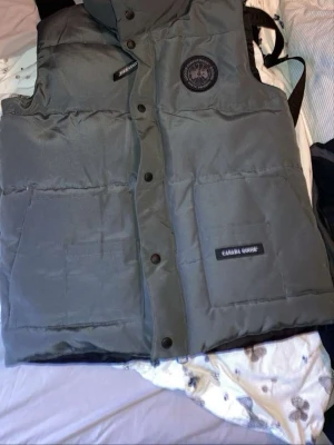Grå dunväst från Canada Goose - Snygg grå dunväst från Canada Goose med svart logotyp på bröstet och två stora fickor framtill. Västen har hög krage, tryckknappar och klassisk Canada Goose-märkning. Perfekt för lager-på-lager och håller dig varm under kyliga dagar.