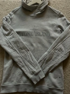 Ljusgrå hoodie från Stone Island - Snygg ljusgrå hoodie från Stone Island med broderad logga på bröstet. Klassisk modell med huva och långa ärmar. Tillverkad i mjukt bomullsmaterial som är skönt mot huden. Perfekt för en avslappnad och trendig stil.