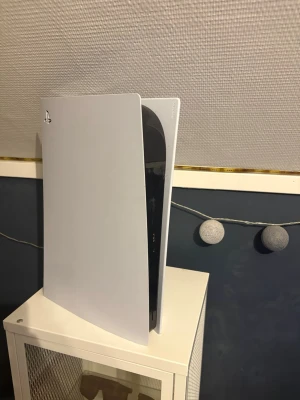 PlayStation 5 konsol från Sony - Säljer en PlayStation 5 konsol från Sony. Konsolen har en futuristisk design med vita och svarta paneler och blå LED-detaljer. Perfekt för gaming med kraftfull prestanda och stöd för de senaste spelen. Står stabilt vertikalt och har USB-portar på framsidan. Man får med hörlurar och kontroll till ps5! 