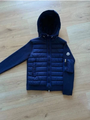 Mörkblå pufferjacka från Moncler - Snygg mörkblå pufferjacka från Moncler med huva och dragkedja framtill. Jackan har quiltad front och huva i glansigt material, medan ärmarna och ryggen är i mjukare tyg. Ficka med dragkedja och Moncler-logga på ena ärmen. Perfekt för kalla dagar.