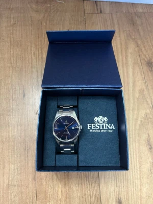 Festina silverfärgad klocka i stål - Festina herrklocka med metallarmband och blå urtavla. Klockan levereras i originalask från Festina. I asken finns extra länkbitar för att kunna justera armbandet efter handledens storlek. Klassisk analog modell med tim- och sekundvisare. Passar som vardags- och dressklocka för herr.  Observera att två ”av siffrorna har lossnat. Men det är lätt fixat om man är händig. Även en liten repa i glaset.  Annars i fint skick! Pris kan diskuteras!  /Mvh Lukas