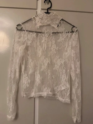 Vit spetsblus med hög krage - Säljer en superfin vit spetsblus med lång ärm och hög, rynkad krage. Blusen är transparent med blommigt spetsmönster och har en liten knytning framtill vid halsen. Perfekt för dig som gillar romantisk och trendig stil.