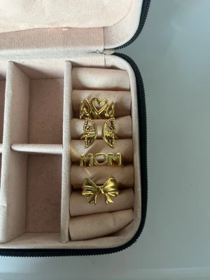 Fyra guldiga ringar med motiv - Säljer ett set med fyra guldiga ringar i olika designer: en med hjärtan runt om, en med rosett, en med texten 'MOM' och en med fjärilsvingar. Alla ringar har en modern och trendig look, perfekta för att mixa och matcha. Materialet är metall i guldfärg. Fjärilen samt den med hjärtan har jag köpte från små företag och dem andra två e köpta från shein💕