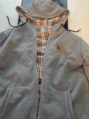 Grå zip-hoodie från Burberry - Grå hoodie med dragkedja från Burberry. Klassisk huva med dragsko och ikoniskt rutigt foder på insidan. Broderad logga på bröstet och BURBERRY-tryck baktill vid midjan. Mjuk bomullsblandning och två fickor framtill.