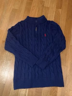 Mörkblå kabelstickad tröja Polo Ralph Lauren - Mörkblå kabelstickad tröja från Polo Ralph Lauren med klassisk röd logga broderad på bröstet. Tröjan har rund halsringning, långa ärmar och ribbade muddar. Perfekt för dig som gillar tidlös och stilren design. Den är lite tightare på ärmarna och mindre än M. Passar ca 12-14 åring.