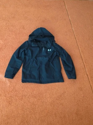 Under armour windbreaker - Svart vindjacka från Under Armour med huva och halv dragkedja framtill. Jackan har en sportig look och är tillverkad i ett lätt, vattenavvisande material. Perfekt för blåsiga dagar och outdoor-aktiviteter.