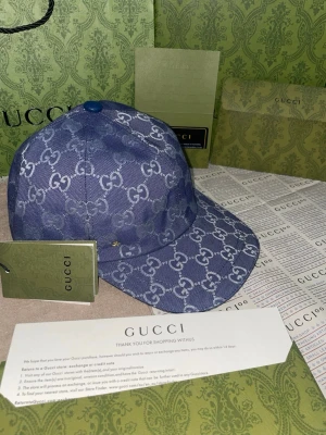Blå keps från Gucci med monogram - Snygg blå keps från Gucci med klassiskt GG-monogram i vitt över hela kepsen. Justerbar rem baktill i blått läder med präglad logotyp. Perfekt accessoar för dig som vill sticka ut med exklusiv streetstyle.