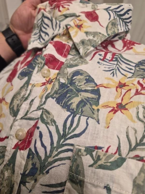 Blommig skjorta med färgglatt mönster Dressmann linnen blend L - Snygg skjorta i linne med blommigt mönster i rött, gult, grönt och blått på vit botten. Skjortan har klassisk krage, knappar framtill och lång ärm. Perfekt för dig som vill sticka ut med en färgglad och somrig stil.
