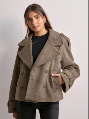 Beige jacka med bred krage - Stilren beige jacka med dubbelknäppning, bred krage och axelklaffar. Jackan har två framfickor och dekorativa knappar vid ärmsluten. Tillverkad i ett ull-liknande material som ger en klassisk look.