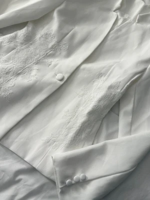 Vit kavaj med spetsdetaljer - Stilren vit kavaj med eleganta spetsbroderier på framsidan. Kavajen har en klädda knapp framtill och tre dekorativa knappar vid ärmslutet. Skräddad passform och mjukt tyg ger en lyxig känsla. Perfekt för dig som vill sticka ut med en unik och trendig look.