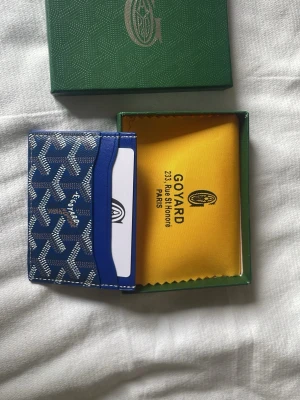 Blå korthållare från G - Snygg blå korthållare från G med deras ikoniska mönster i vitt och grått. Korthållaren har flera fack för kort och är tillverkad i läder med canvasdetaljer. Perfekt för dig som vill ha en stilren och exklusiv accessoar.