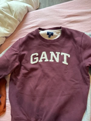 Vinröd sweatshirt från GANT - Säljer en vinröd sweatshirt från GANT med vit broderad logga på bröstet. Tröjan har rund halsringning och ribbade muddar vid ärmslut och nederkant. Perfekt för en avslappnad och stilren look. Pasaar s/m