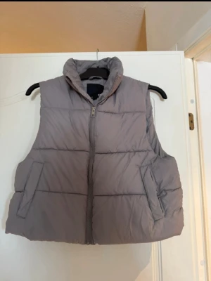 Grå dunväst från 157 - Snygg grå dunväst från 157 med hög krage och dragkedja framtill. Västen har två snedställda fickor och quiltad design som ger en puffig look. Perfekt att slänga över en hoodie eller tröja för extra värme.