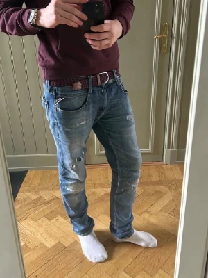 Replay ambass slitningar - Säljer ett par blå jeans med slitningar och distressed detaljer på benen. Jeansen har en slim fit passform och klassiska fem fickor. Materialet är denim och färgen är ljusblå med vita och grå nyanser på slitningarna. Perfekt för dig som gillar en avslappnad och trendig look. L32 w31