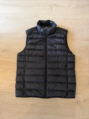 Svart dunväst från Uniqlo - Snygg svart dunväst från Uniqlo med quiltad design och hög krage. Västen har dragkedja framtill och är lättviktig, perfekt att slänga över en hoodie eller tröja för extra värme. Enkel och stilren look som funkar till det mesta.