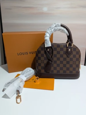 Iouis Vuitton Alma BB. - En tidlös famn av elegans Denna vackra skapelse vilar med en naturlig värdighet, klädd i ett djupt och harmoniskt schackrutigt mönster i varma toner av choklad och ebenholts. Dess mjuka, rundade former viskar om en klassisk skönhet som aldrig går ur tiden, en trofast följeslagare som bär dina hemligheter med stil.