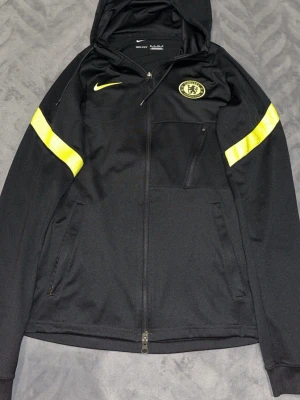 Chelsea Nike svart hoodie Dri-FIT - Snygg svart hoodie från Nike med Chelsea FC-logga på bröstet och gula detaljer på ärmarna. Tillverkad i Dri-FIT-material som andas, perfekt för träning eller chill. Dragkedja framtill, fickor med dragkedja och huva.
