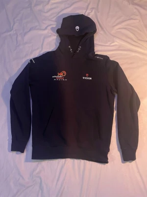 Alinghi Red Bull Racing hoodie - Mörkblå hoodie med tryck från Alinghi Red Bull Racing och Tudor på bröstet. På huvan ser du också ett klassiskt sail racing märke. Perfekt för dig som gillar racing och sportig stil perfekt till träning.