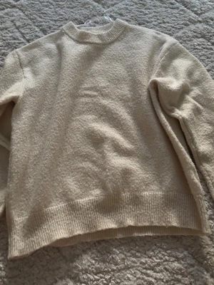 Beige stickad tröja med rund hals - Mysig beige stickad tröja med rund halsringning och ribbade muddar vid ärmslut och nederkant. Perfekt för lager på lager och enkel att matcha med olika outfits. Tröjan har en avslappnad passform och är tillverkad i ett mjukt material.