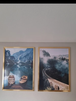 Två tavlor från kända Desenio med naturmotiv i guldram - Desenio-tavla i svart/beige toner, med guldiga ram i storlek 70×100 cm. Modern och stilren. Mycket fint skick. två snygga tavlor med naturmotiv, båda i guldfärgade ramar. Ena tavlan visar två träbåtar vid en spegelblank sjö med berg i bakgrunden, den andra ett dimmigt landskap med en kurvig stenbro. Perfekta som väggdekoration för dig som gillar natur och äventyr.
