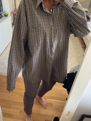 Rutig oversized skjorta från CBC - Rutig skjorta från CBC i bruna och beige toner. Oversized passform med klassisk krage, bröstficka och knäppning framtill. Perfekt att styla lager på lager. Skön och luftig känsla, troligtvis i bomull.
