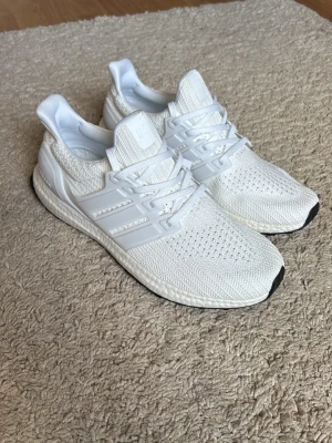 Vita Adidas Ultraboost sneakers - Snygga helvita Adidas Ultraboost sneakers med stickad ovandel i mesh och klassiska tre ränder på sidan. Skorna har en bekväm Boost-sula och svarta detaljer på sulans framdel. Perfekta för dig som gillar stilrena och sportiga sneakers.