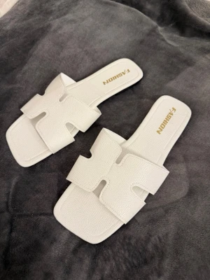Vita slip-in sandaler från Fashion - Stilrena vita sandaler i slip-in modell från Fashion. De har breda band med ett utstansat H-mönster och fyrkantig tå. Dem är aldrig använda. Tillverkade i ett konstläder med lätt textur för en trendig look. Perfekta för en avslappnad och modern stil. 