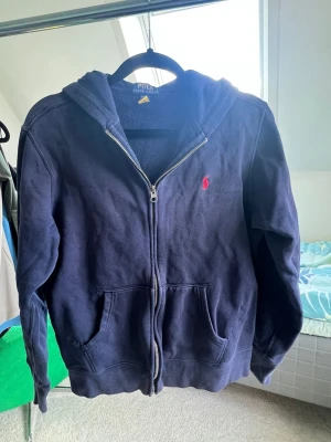Mörkblå hoodie från Polo Ralph Lauren - Mörkblå hoodie med dragkedja från Polo Ralph Lauren. Klassisk design med huva, kängurufickor och röd broderad logga på bröstet.
