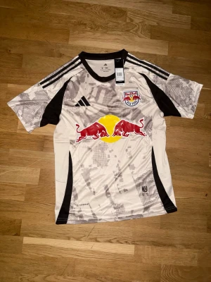 NY Red Bulls matchtröja Adidas - Säljer en vit och svart matchtröja från Adidas för New York Red Bulls med klubbmärke och Red Bull-logga på bröstet. Tröjan har korta ärmar, rund hals och detaljer i grått samt svarta ränder på axlarna. Perfekt för fotbollsträning eller support.
