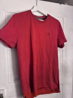 Röd t-shirt från Superdry Athletics - Snygg röd t-shirt från Superdry Athletics med diskret broderad logga på bröstet. Klassisk rund halsringning och korta ärmar. Tillverkad i mjuk bomull som är skön att bära hela dagen. Passar snarare M.