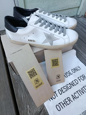 Golden Goose vita sneakers med stjärna - Golden Goose sneakers i vitt läder med grå mockadetaljer och silverfärgad stjärna på sidan. Skorna har grå skosnören, svart hälkappa och klassisk låg siluett. Snygg och trendig design med vintagekänsla och ikoniska detaljer.