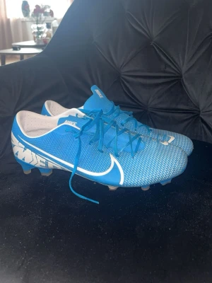 Blå Nike Mercurial fotbollsskor - Säljer ett par blå Nike Mercurial fotbollsskor med vita detaljer och stor vit swoosh på sidan. Skorna har snörning, låg profil och dobbar för spel på gräs. Ovandelen är i syntetmaterial för bra bollkontroll och passform. Perfekta för dig som vill sticka ut på planen!