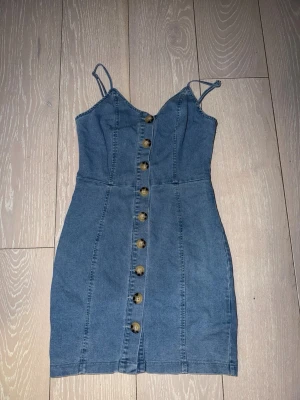 Blå jeansklänning med knappar - Snygg blå jeansklänning med smala axelband och knappar hela vägen framtill. Klänningen är figurnära och har en kort längd, perfekt för en trendig look. Tillverkad i jeansmaterial som ger en cool och avslappnad vibe.