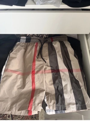 Burberry shorts - Snygga shorts i beige med klassiskt rutigt mönster i svart, vitt och rött. Shortsen har elastisk midja med snörning och en bakficka. Perfekta för sommaren och har en avslappnad passform.