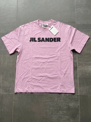 Rosa Jil Sander t-shirt i bomull - Snygg rosa t-shirt från Jil Sander med svart logotyptryck på bröstet. Klassisk passform med rund hals och korta ärmar. Tillverkad i 100% bomull för en mjuk och skön känsla. Perfekt för en stilren och trendig look.