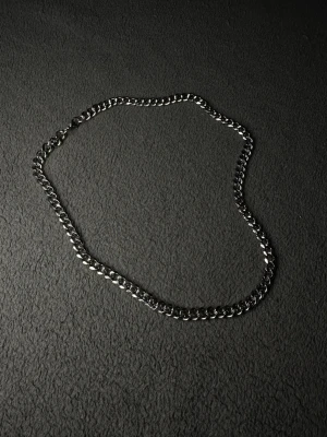 Cubansk Kedja i Silverfärg 50cm 7mm – Rostfritt Stål  - 50cm långt, 7mm bred. Stilrent halsband med breda, platta länkar i silverfärgad metall. Kedjan har en klassisk design och stängs med ett karbinlås. Perfekt för att ge din outfit en cool och modern touch.