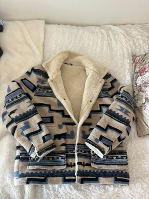Beige teddyjacka med blått mönster - Snygg teddyjacka i beige med blått och svart geometriskt mönster. Jackan har stor krage, två stora fickor framtill och mysigt teddyfoder. Perfekt för kalla dagar när du vill sticka ut lite extra.