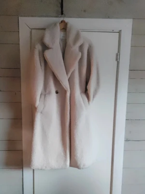 Lång fluffig teddy-kappa i beige - Säljer en supermjuk och lång teddy-kappa i beige med breda slag och fickor på sidorna. Kappan har en oversized passform och stängs med en knapp framtill. Perfekt för kalla dagar när du vill vara både varm och snygg.