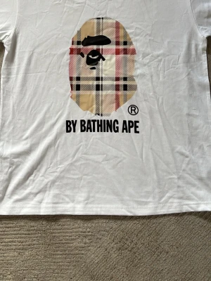 Vit BAPE t-shirt med rutig logga - Vit t-shirt från BAPE med den klassiska apansiluetten i ett rutigt mönster på bröstet och texten 'BY BATHING APE' under. T-shirten har en avslappnad passform och är tillverkad i mjuk bomull. Perfekt för dig som gillar streetwear och vill sticka ut.