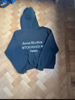 Svart hoodie Acne Studios Stockholm - Svart hoodie från Acne Studios med stor huva och tryck på ryggen där det står 'Acne Studios STOCKHOLM 1996'. Klassisk passform med ribbade muddar vid ärmslut och midja. Perfekt för en avslappnad och trendig stil.